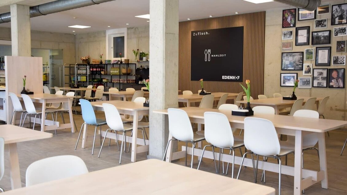 Ein modernes Restaurant mit hellen Holzmöbeln und weißen Stühlen. An der Wand hängen zahlreiche Bilder, und im Hintergrund ist eine Theke sichtbar. | © Edenhof