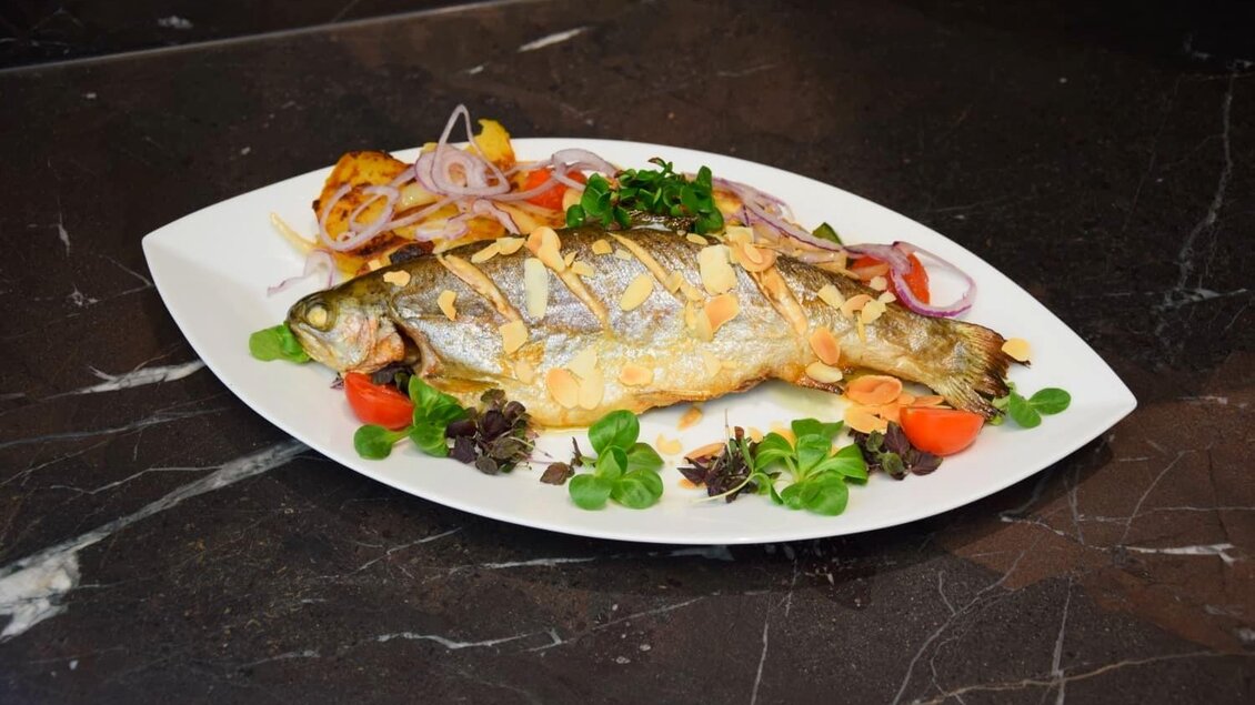 Ein gebratener Fisch auf einem weißen Teller, garniert mit frischen Kräutern, Zwiebeln und Tomaten. Daneben liegen knusprige Kartoffelstreifen. | © Edenhof