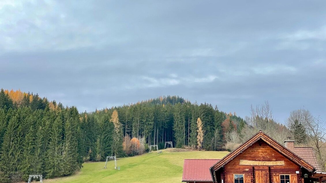 Eine charmante Holzhütte steht in einer grünen Landschaft. Im Hintergrund sind Wälder und eine sanfte Anhöhe zu sehen. | © Marcus Auer