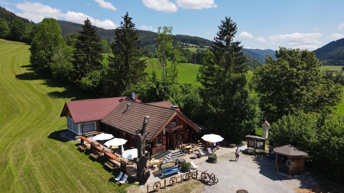 Eine gemütliche Hütte umgeben von grünen Wiesen und hohen Bäumen. Tische und Sonnenschirme im Freien laden zum Entspannen ein. | © Marcus Auer