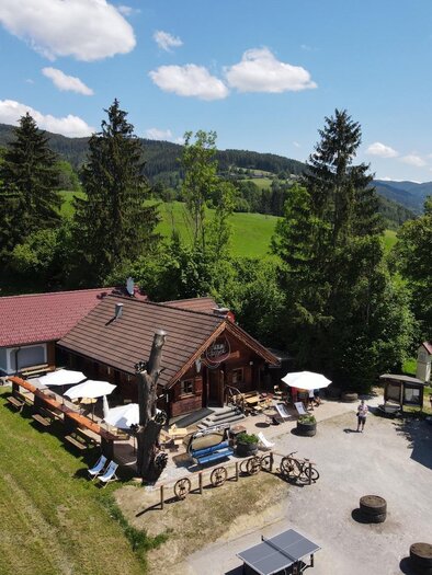 Eine gemütliche Hütte umgeben von grünen Wiesen und hohen Bäumen. Tische und Sonnenschirme im Freien laden zum Entspannen ein. | © Marcus Auer