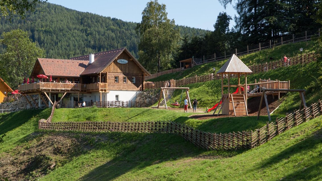 Ein hübsches Holzhaus steht auf einem grünen Hügel, umgeben von Bäumen. Im Vordergrund befindet sich ein Spielplatz mit Rutschen und Schaukeln. | © TV Hochsteiermark
