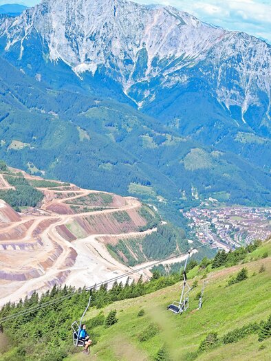 A picturesque mountain view with a large mountain range in the background. In the foreground, there are cable cars passing over a green meadow. | © Viktoria Hirtler