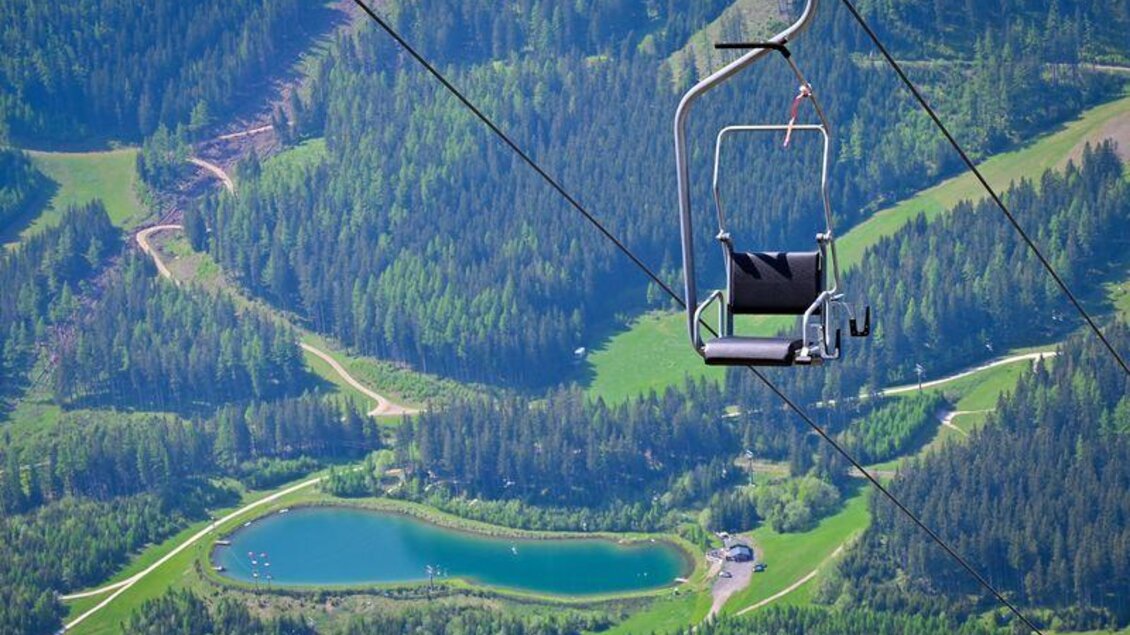 Eine Bergbahn schwebt über saftige Wiesen und einen kleinen See. Darunter erstreckt sich eine grüne Landschaft mit Wäldern. | © Präbichl Bergbahnen