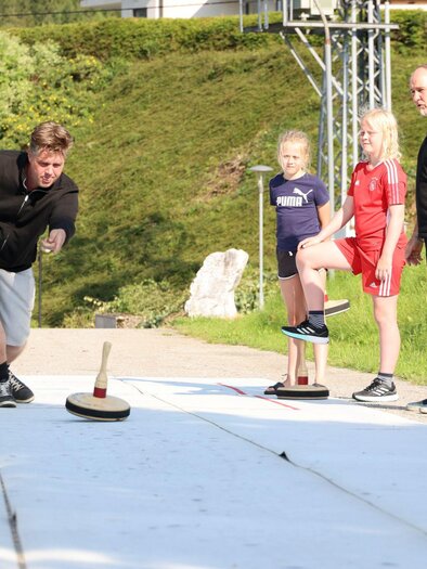 Eine Gruppe von Menschen spielt draußen eine Sportart. Im Vordergrund wirft ein Mann einen Klinker, während zwei Mädchen und ein weiteres Paar zuschaut. | © Stephan Steiner
