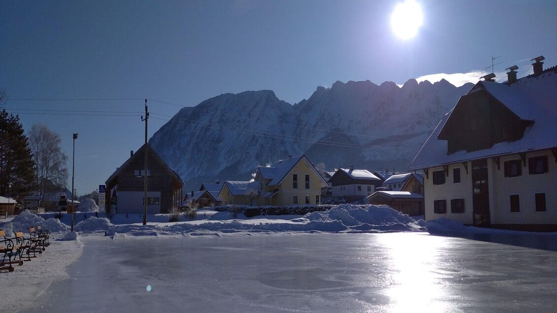 Eine winterliche Landschaft mit schneebedeckten Bergen und einem gefrorenen Teich. Die Sonne scheint hell über den ruhigen Häusern. | © Johanna Provatopoulos