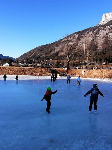 Eine Eislauffläche mit vielen Menschen, die auf dem Eis stehen und skaten. Im Hintergrund sind Berge und ein klarer blauer Himmel zu sehen. | © René Haselnus