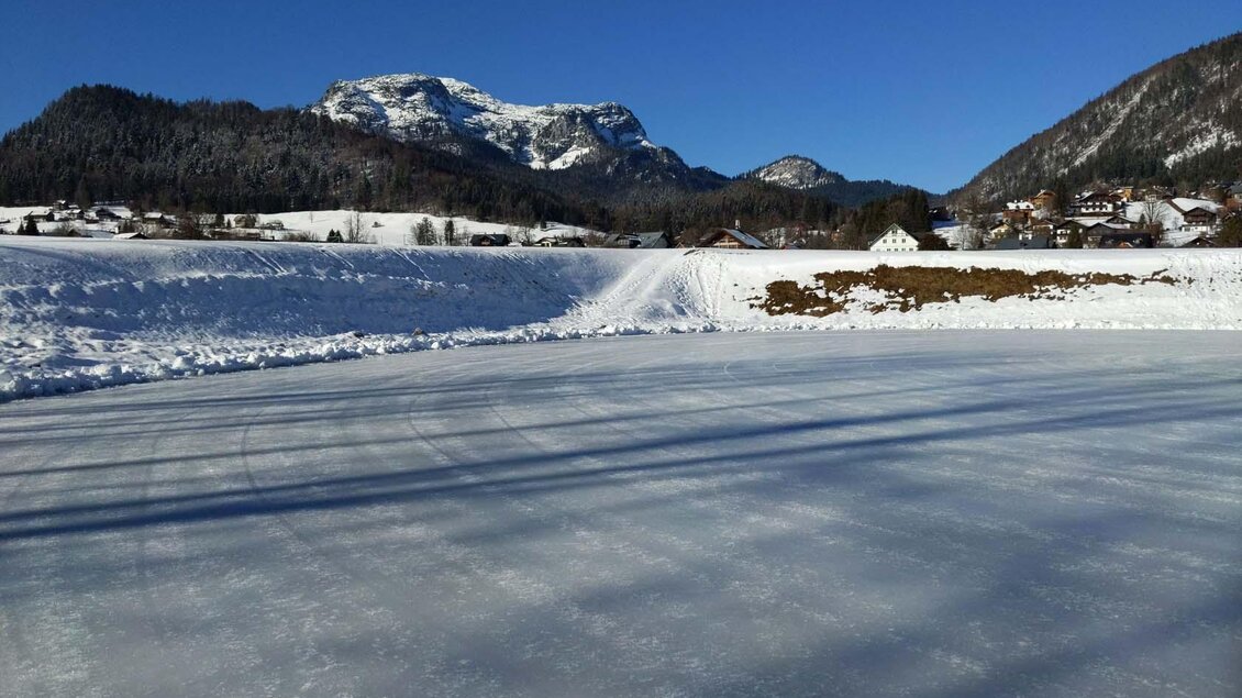Eine gefrorene Fläche mit Schnee und Eis, umgeben von Bergen und einem klaren blauen Himmel. Im Hintergrund sind einige Häuser sichtbar. | © René Haselnus