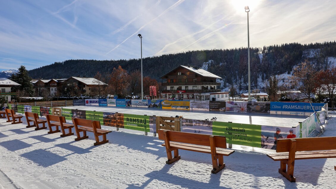 Eine Eislaufbahn in einem winterlichen Dorf mit schneebedeckten Bänken. Die Umgebung ist von Bergen und einem klaren Himmel umgeben. | © RVB Ramsau