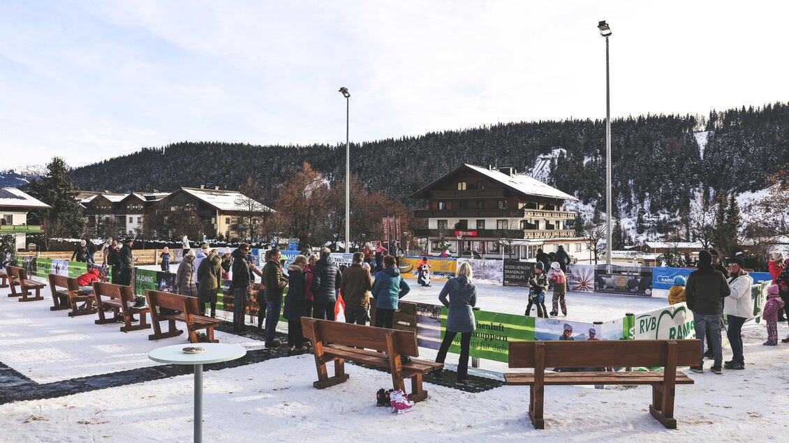Eine belebte Eislaufbahn in einer winterlichen Landschaft. Menschen genießen das Eislaufen, umgeben von schneebedeckten Bergen und gemütlichen Hütten. | © RVB Ramsau