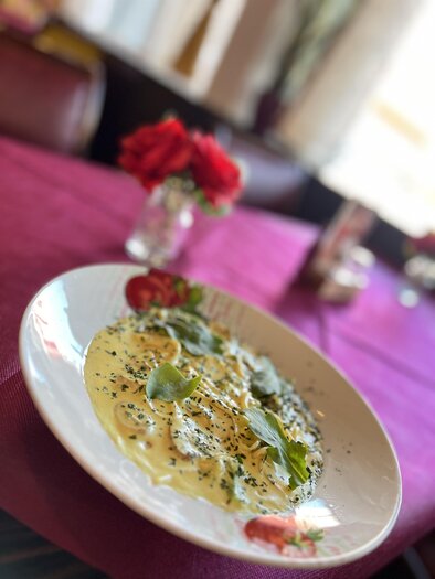 Ein Teller Pasta mit einer cremigen Sauce und frischen Kräutern. Im Hintergrund sind Blumen und ein gedeckter Tisch sichtbar.