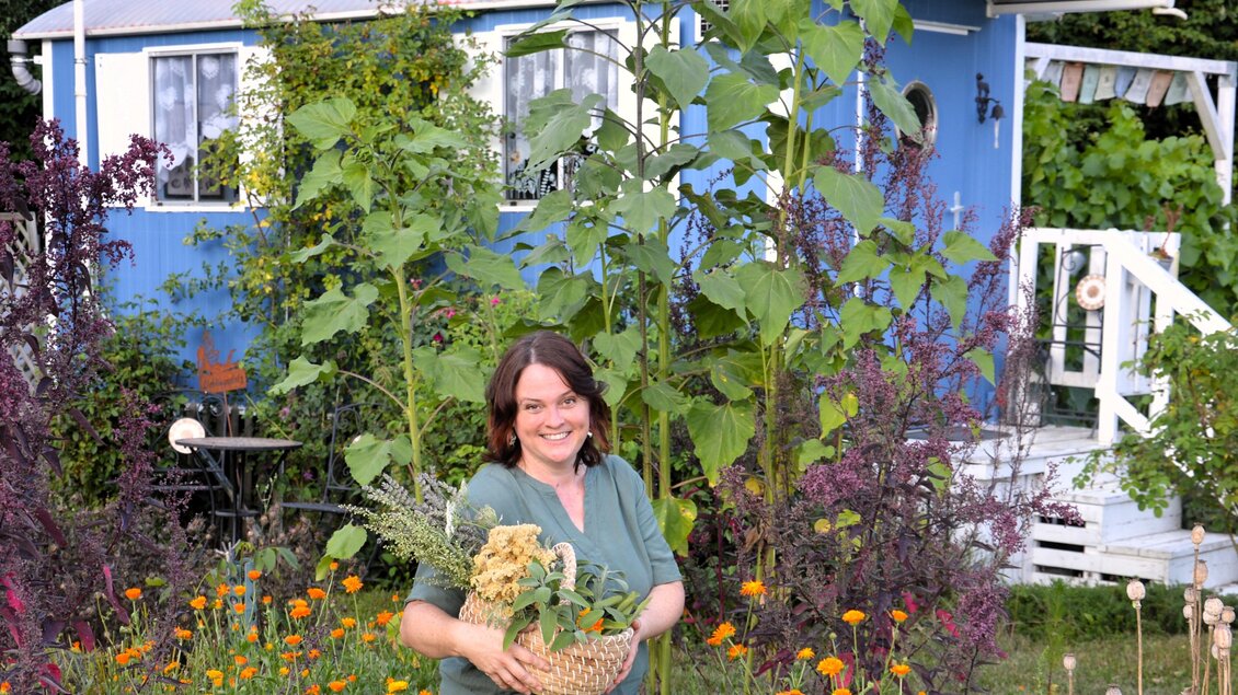 Eine lächelnde Frau steht in einem bunten Garten. Im Hintergrund sieht man ein blaues Haus und hohe Sonnenblumen. | © Martina Schmidt
