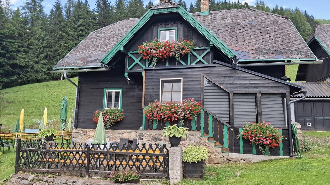 Ein gemütliches Holzhaus mit bunten Blumenkästen und einem gepflegten Garten. Umgeben von grünen Wiesen und Bäumen, strahlt es ländlichen Charme aus. | © Johanna Elsner