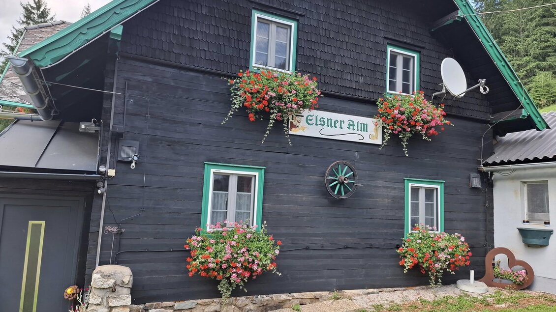 Ein traditionelles Holzhaus mit einer dunklen Fassade und grünen Details. Es sind bunte Blumenkästen an den Fenstern angebracht. | © Johanna Elsner