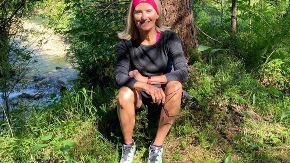 Eine Frau sitzt im Grünen, umgeben von Bäumen und einem kleinen Fluss im Hintergrund. Sie trägt sportliche Kleidung und lächelt freundlich. | © Manuela Schrittwieser