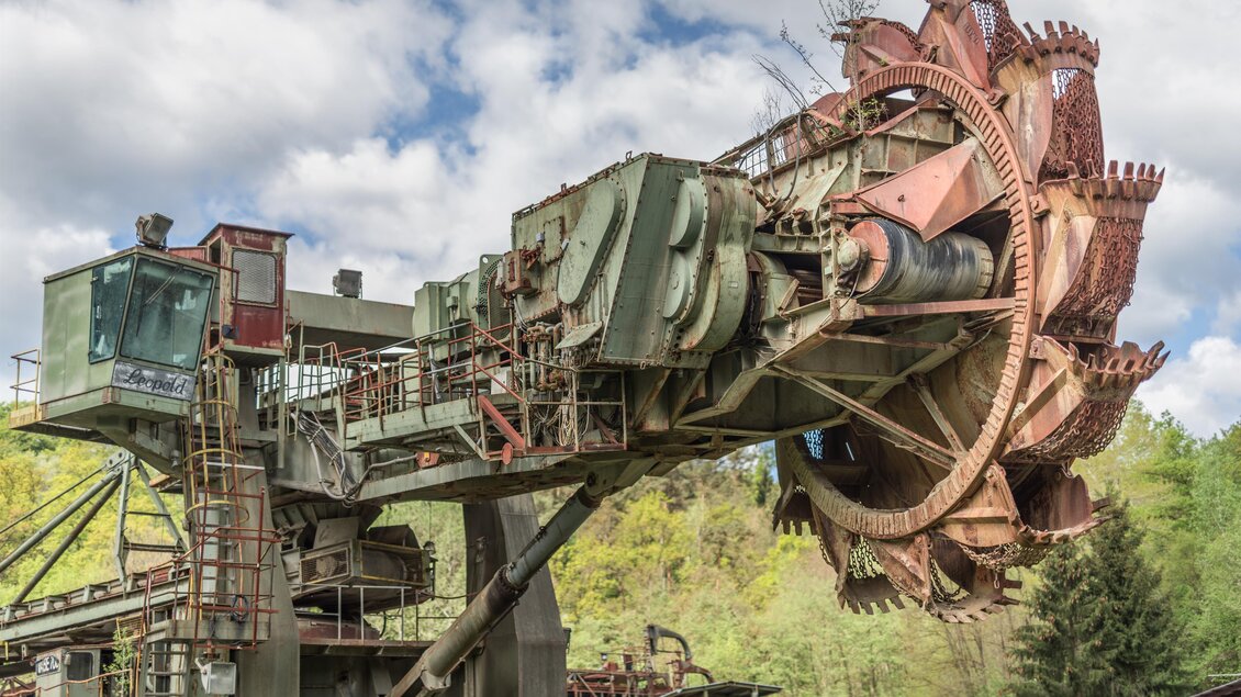 Eine große, industrielle Fördermaschine mit einem kreisförmigen Schneidwerkzeug. Im Hintergrund sind Bäume und ein bewölkter Himmel sichtbar. | © Lipizzanerheimat - Die Abbilderei