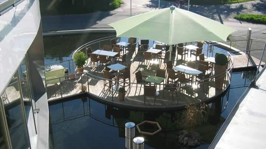 Terrasse mit Sonnenschirm und Sitzplätzen am künstlich angelegten Wasser beim Energieschiff Mochart in Köflach. | © Mochart GmbH