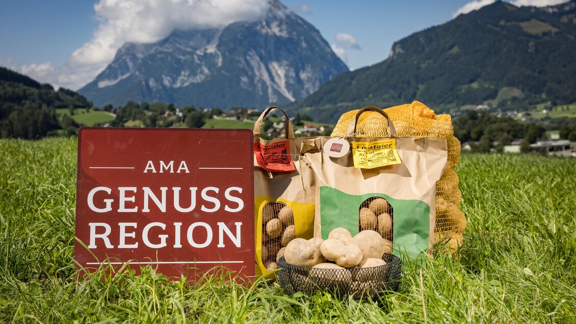 Ein Schild mit der Aufschrift „AMA Genuss Region“ steht in einer grünen Wiese. Im Vordergrund befinden sich Obst- und Gemüsebeutel mit Kartoffeln und im Hintergrund sind majestätische Berge zu sehen. | © Netzwerk Kulinarik Wildbild