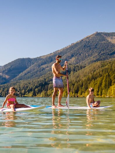 Eine Familie steht auf Stand-Up Paddle-Boards im ruhigen Wasser eines Sees. Im Hintergrund sind Berge und Bäume zu sehen. | © TV Hochsteiermark | Rudy Dellinger