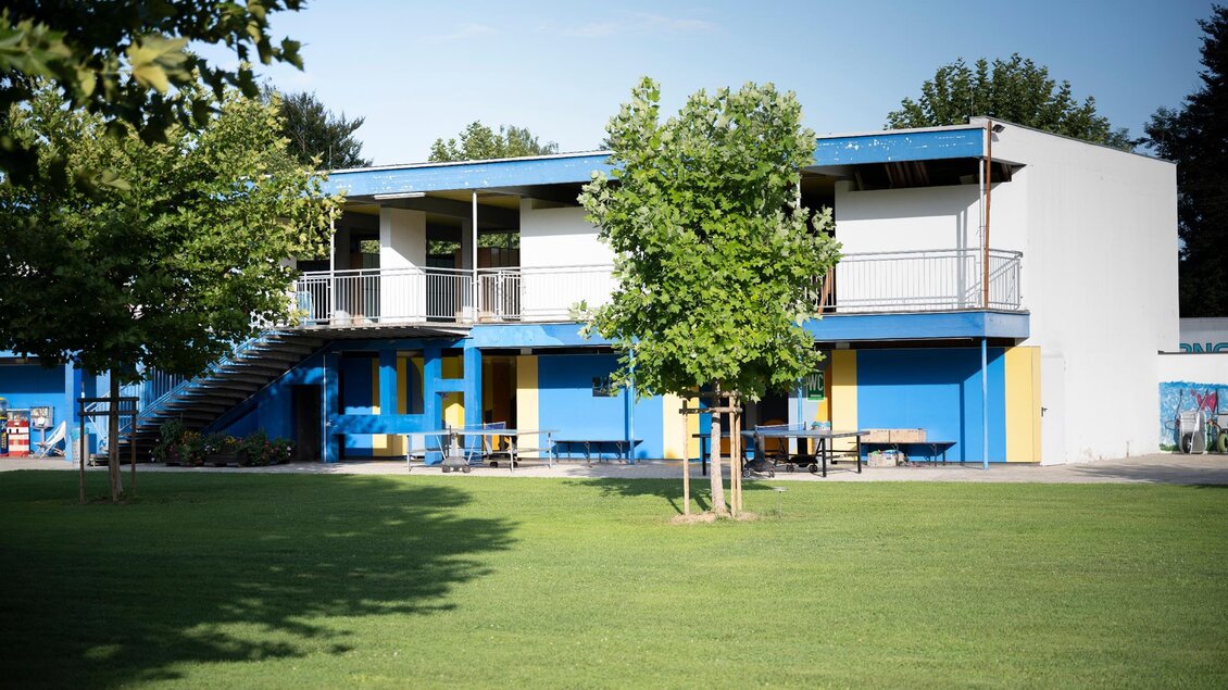 Ein modernes Gebäude mit blauer und gelber Fassade steht in einem grünen Rasenbereich. Einige Bäume umgeben den Bereich und es gibt Möbel im Freien. | © Schilcherland Steiermark