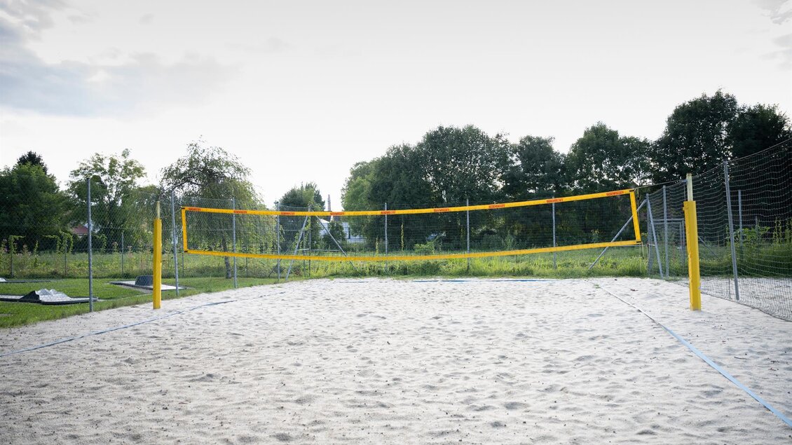 Ein Volleyballfeld aus Sand mit einem orangefarbenen Netz. Im Hintergrund sind Bäume und eine Spielplatzfläche sichtbar. | © Schilcherland Steiermark