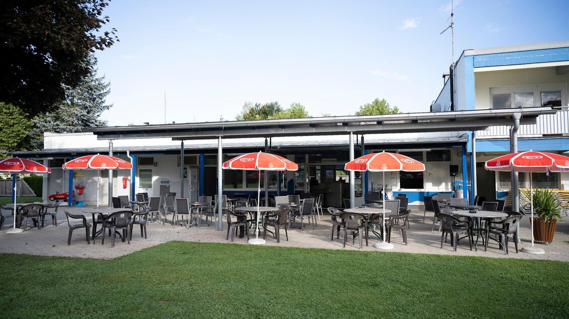 Ein gemütliches Restaurant mit mehreren Tischen und roten Sonnenschirmen im Freien. Im Hintergrund ist ein Gebäude mit einem modernen Design zu sehen. | © Schilcherland Steiermark