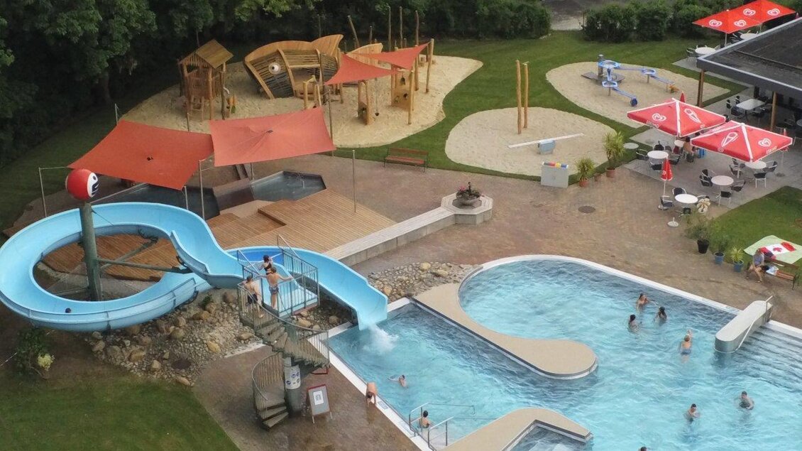 Ein Schwimmbad mit einer Rutsche und einem Spielplatz im Hintergrund. Sonnenschirme und Liegestühle laden zum Entspannen ein. | © Erlebnisbad Pöllau