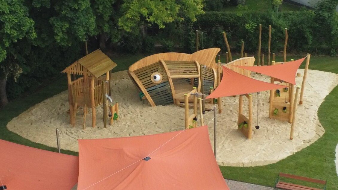Ein Spielplatz mit einer Holzkonstruktion in Form eines Bootes, umgeben von Sand. Markisen bieten Schatten für Kinder, die spielen. | ©  Erlebnisbad Pöllau