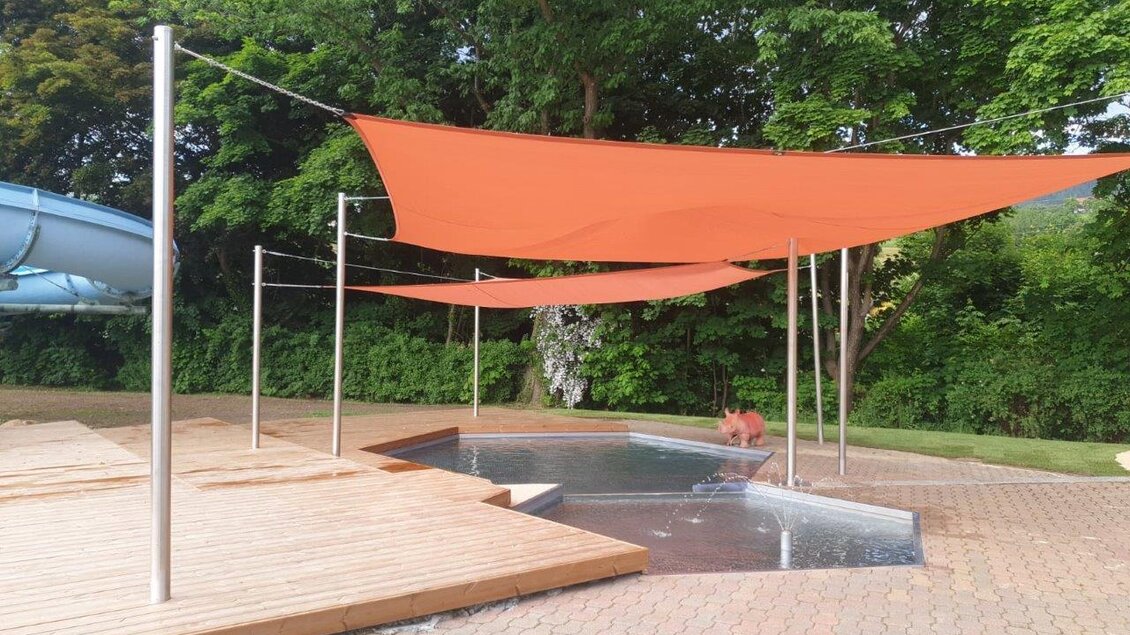Ein Bereich mit orangefarbenen Sonnensegeln über einem kleinen Wasserbecken. Umgeben von Bäumen und einer Holzterrasse, bietet es einen schattigen Platz zum Entspannen. | © Erlebnisbad Pöllau
