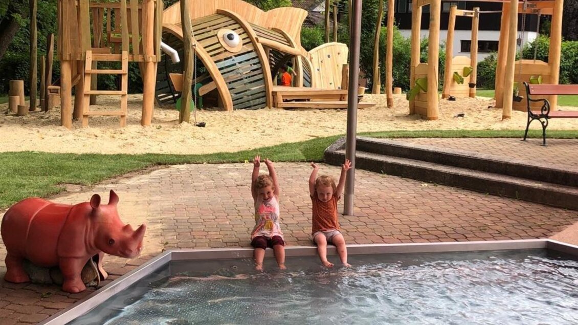 Zwei Kinder sitzen am Ufer eines Pools und halten die Hände hoch. Im Hintergrund sind Spielgeräte und eine große Holzfigur eines Fisches zu sehen. | © Erlebnisbad Pöllau