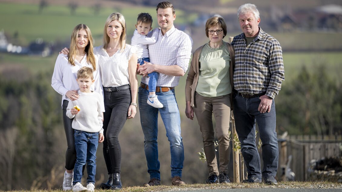 Eine Familie steht im Freien vor einer grünen Landschaft. Sie lächeln und posieren gemeinsam für das Foto. | © König-Kober