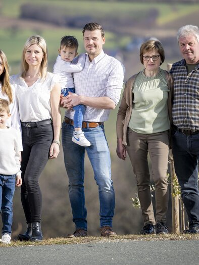 Eine Familie steht im Freien vor einer grünen Landschaft. Sie lächeln und posieren gemeinsam für das Foto. | © König-Kober