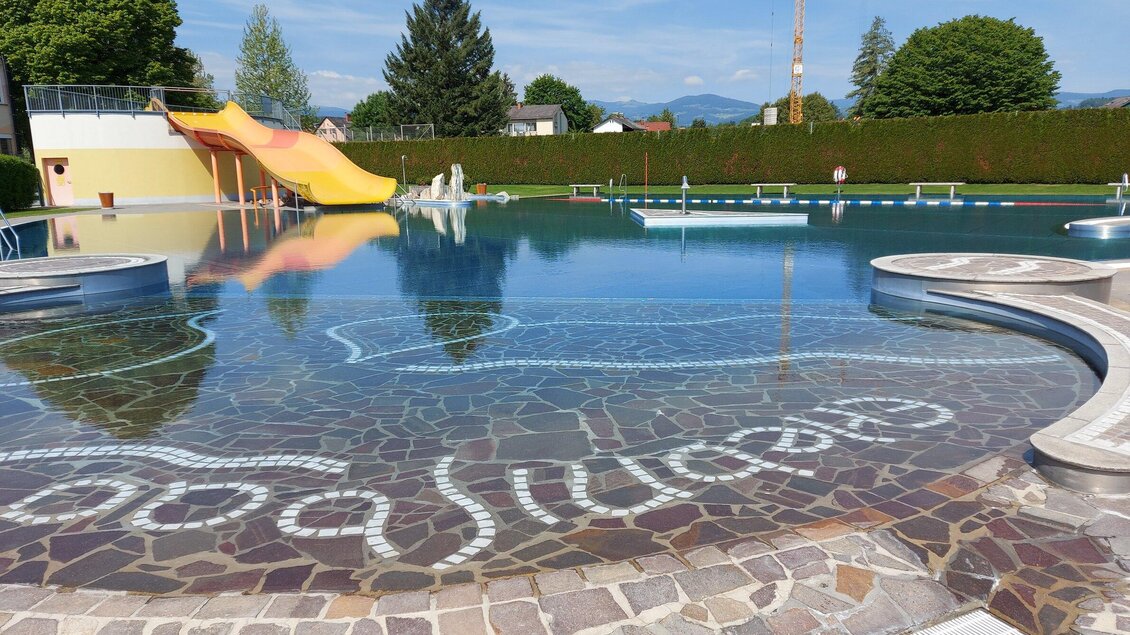 Ein großzügiges Schwimmbad mit einem Rutschenbereich und klarem Wasser. Umgeben von grünen Bäumen und einem blauen Himmel. | © Stadtgemeinde Voitsberg - Mayer
