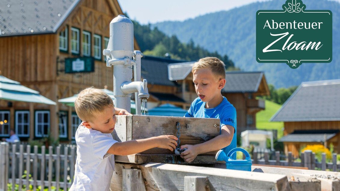 Zwei Kinder spielen im Freien und erkunden eine Holzstruktur. Im Hintergrund sind rustikale Gebäude und eine malerische Landschaft zu sehen. | © www.zloam.at