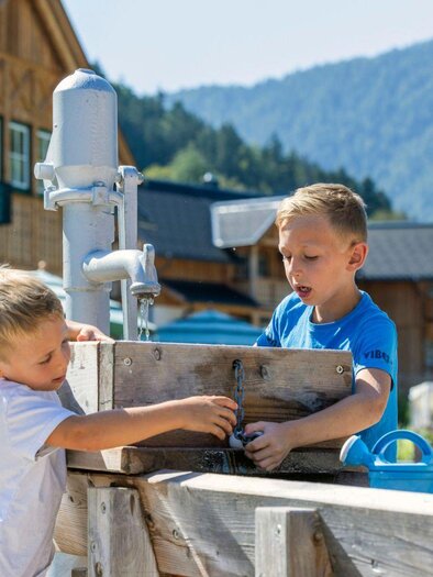 Zwei Kinder spielen im Freien und erkunden eine Holzstruktur. Im Hintergrund sind rustikale Gebäude und eine malerische Landschaft zu sehen. | © www.zloam.at
