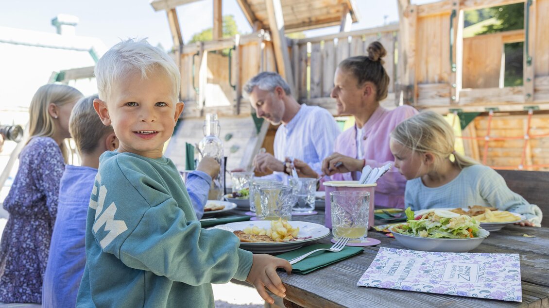 Ein fröhliches Kind sitzt an einem Tisch und lächelt, während eine Familie gemeinsam isst. Der Tisch ist mit Essen und Getränken gedeckt, umgeben von einer Spielumgebung. | © www.zloam.at