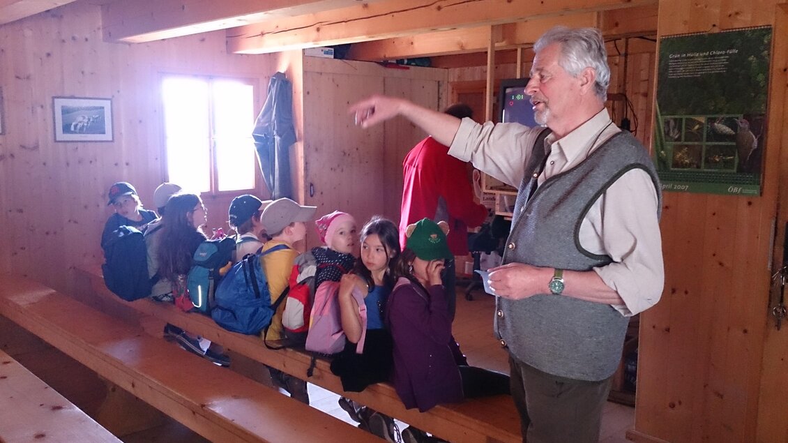 Ein Lehrer erklärt eine Gruppe von Kindern in einem Holzraum. Die Kinder sitzen auf Bankbänken und hören aufmerksam zu. | © TV Hochsteiermark