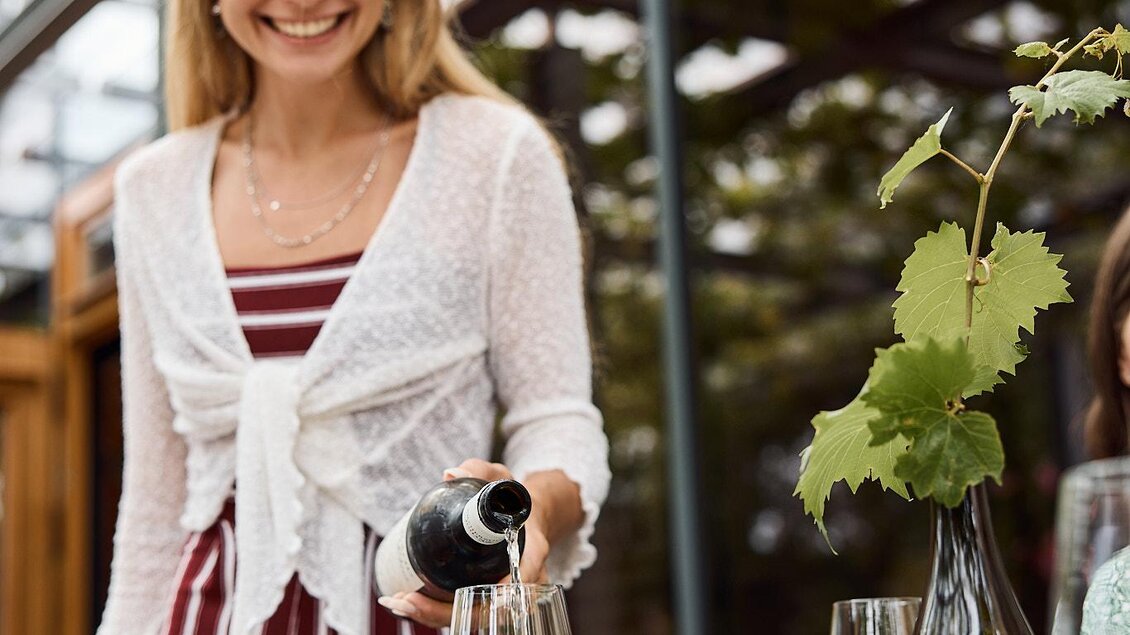 Eine Frau gießt Wein in ein Glas auf einem Tisch. Im Hintergrund sind grüne Pflanzen und ein modernes Gewächshaus zu sehen. | © Erzherzog Johann Weine