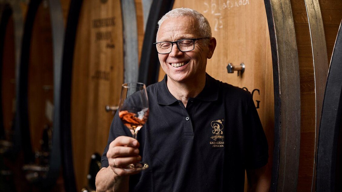 Ein lächelnder Mann hält ein Glas Wein in der Hand. Im Hintergrund sind große Holzfässer zu sehen. | © Erzherzog Johann Weine