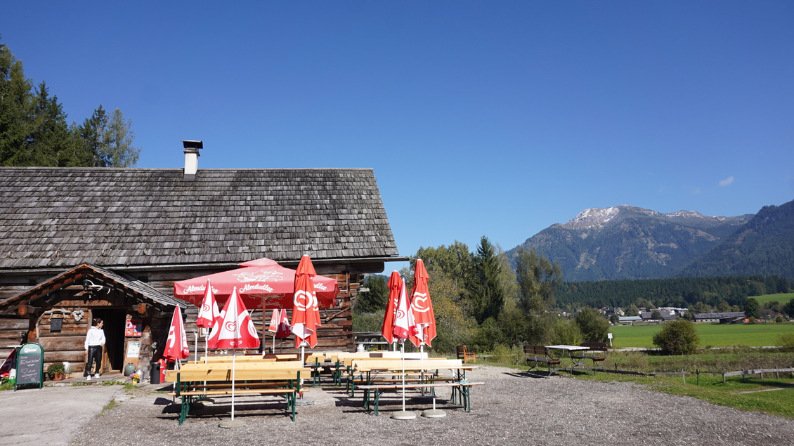 Ein gemütliches Gasthaus in den Bergen mit roten Sonnenschirmen. Im Hintergrund sind grüne Wiesen und Berge zu sehen. | © Eselalm