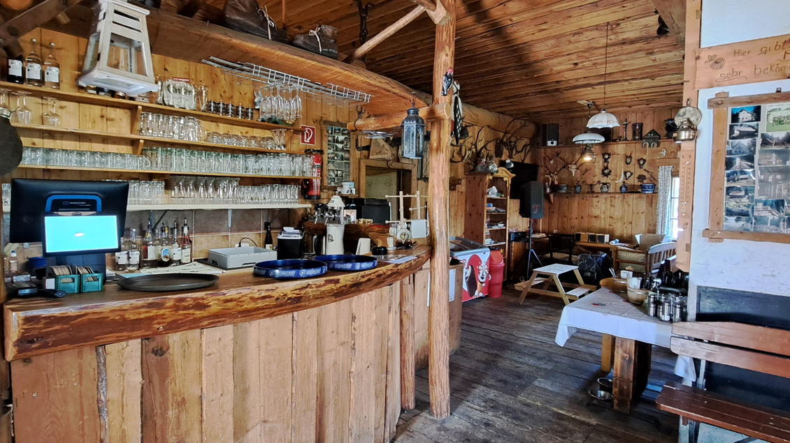 Eine gemütliche Restaurant-Innenansicht mit Holzdekor und einem halb offenen Empfangsbereich. Es gibt Tische für Gäste und an der Bar sind Gläser und Getränke sichtbar. | © Eselalm