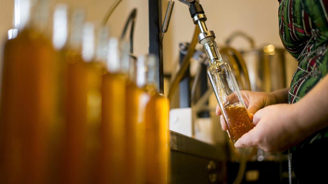 Eine Person füllt mit einer Maschine eine Flasche mit einer goldenen Flüssigkeit. Im Hintergrund sind weitere Flaschen sichtbar. | © Essigmanufaktur Oswald / Schaffer