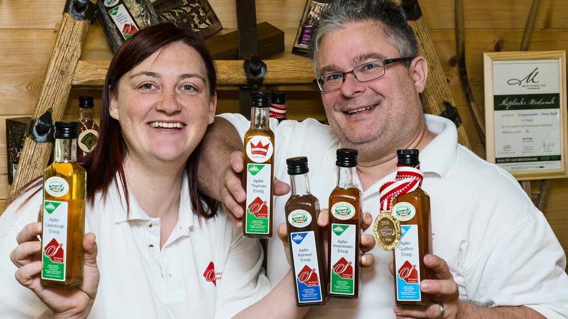 Zwei Personen lächeln und halten Flaschen mit verschiedenen Ölen in der Hand. Im Hintergrund sind Auszeichnungen und Produkte sichtbar. | © Essigmanufaktur