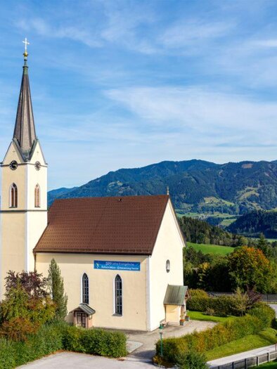 Eine weiße Kirche mit einem hohen Turm steht vor einem klaren blauen Himmel. Umgeben von Bäumen und Hügeln zeigt sie eine ruhige ländliche Szene. | © Evangelische Kirche Gröbming