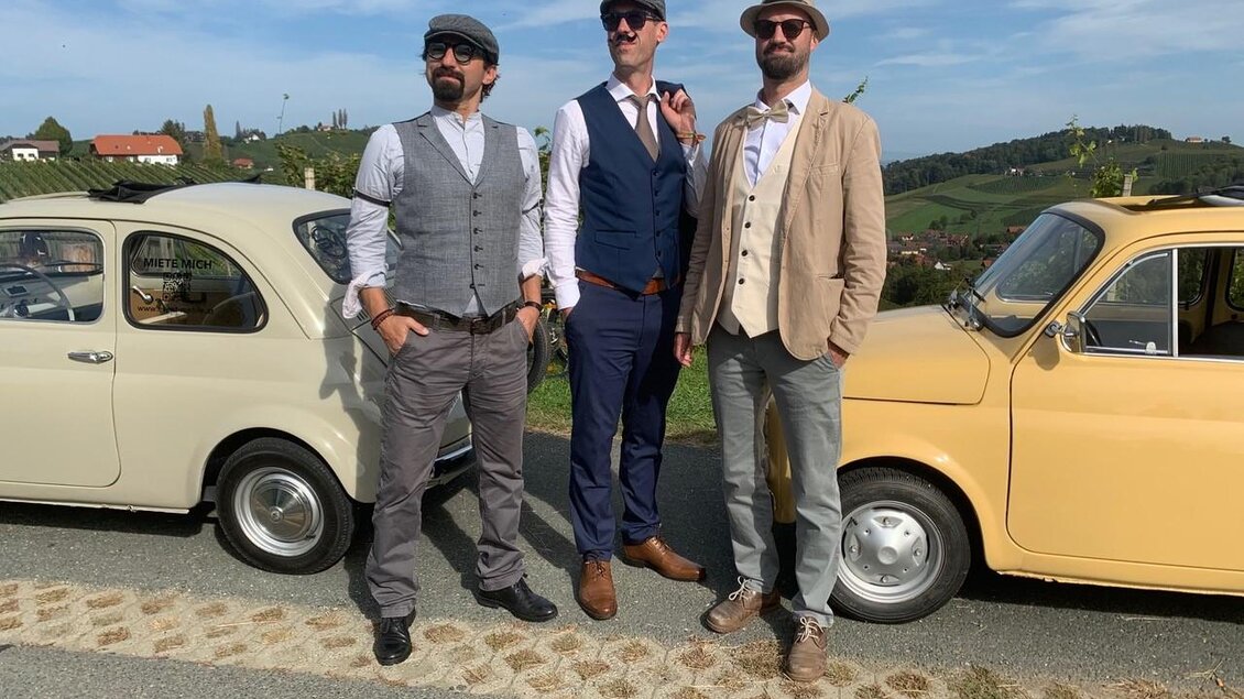 Drei Männer in eleganter Kleidung stehen vor zwei Oldtimern. Im Hintergrund sind grüne Hügel und ein klarer Himmel zu sehen. | © Event Mobile