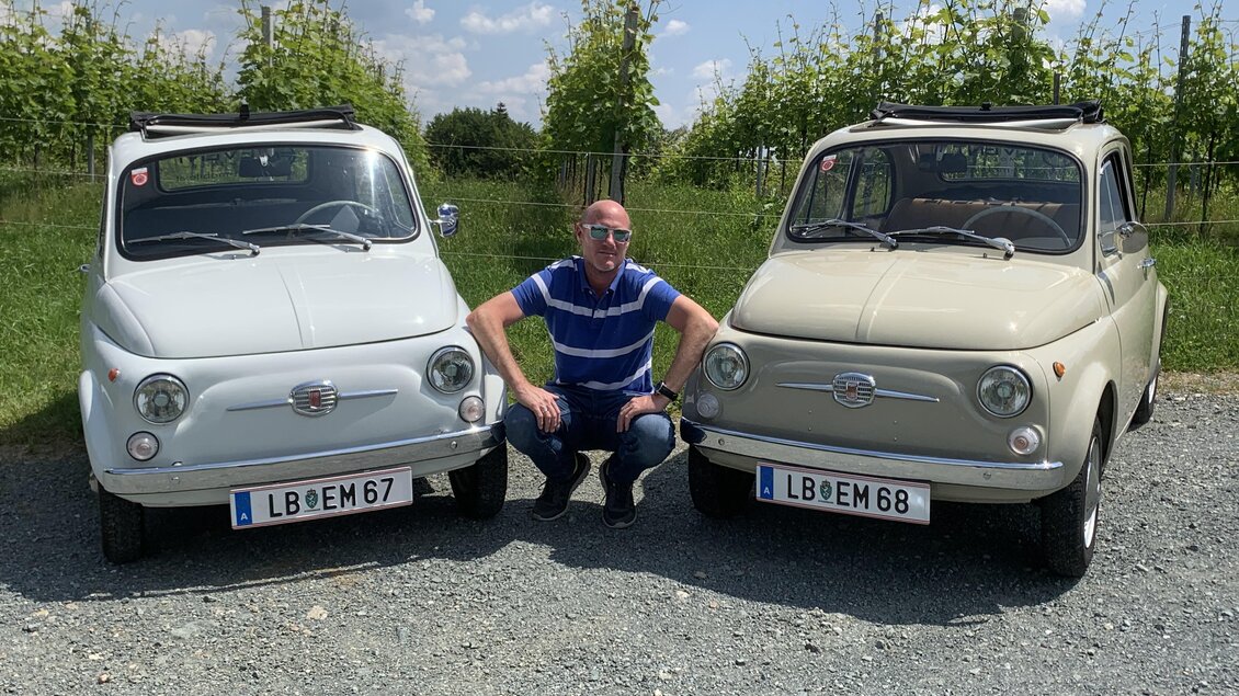 Ein Mann sitzt zwischen zwei klassischen Fiat-Autos auf einem Kiesweg. Im Hintergrund sind Weinreben und blauer Himmel zu sehen. | © Event Mobile