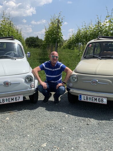 Ein Mann sitzt zwischen zwei klassischen Fiat-Autos auf einem Kiesweg. Im Hintergrund sind Weinreben und blauer Himmel zu sehen. | © Event Mobile