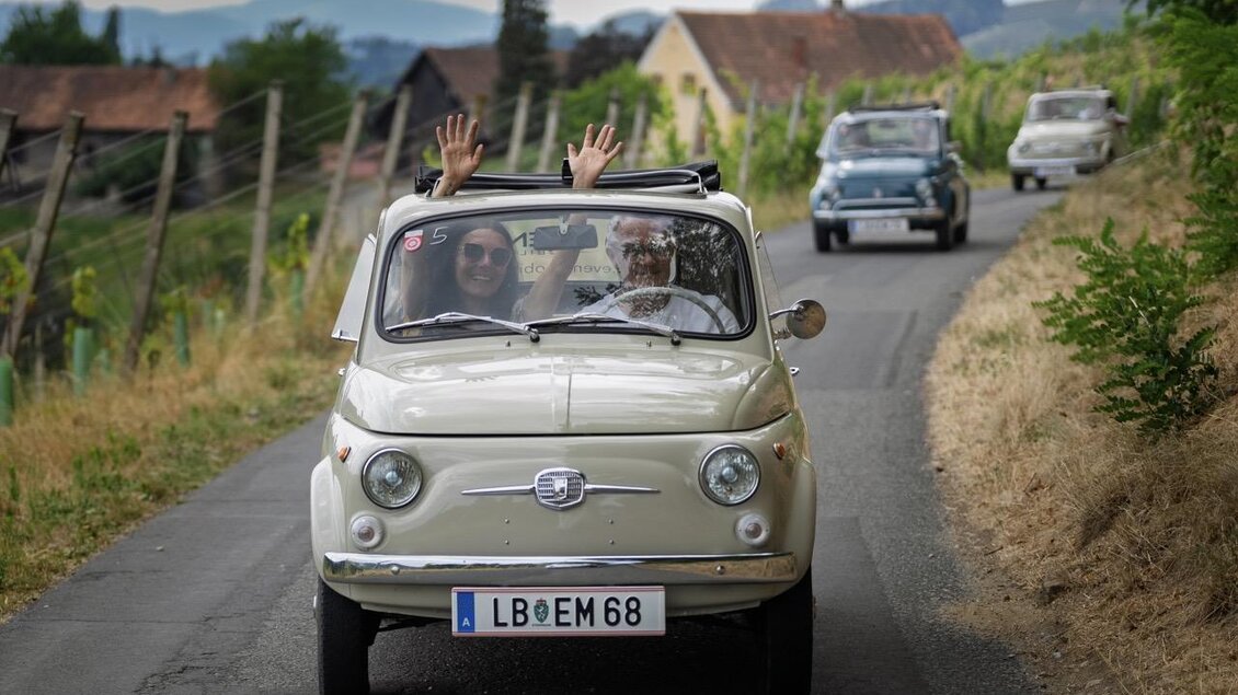 Ein klassisches Auto fährt auf einer ländlichen Straße. Die Fahrerinnen winken fröhlich aus dem Fenster.