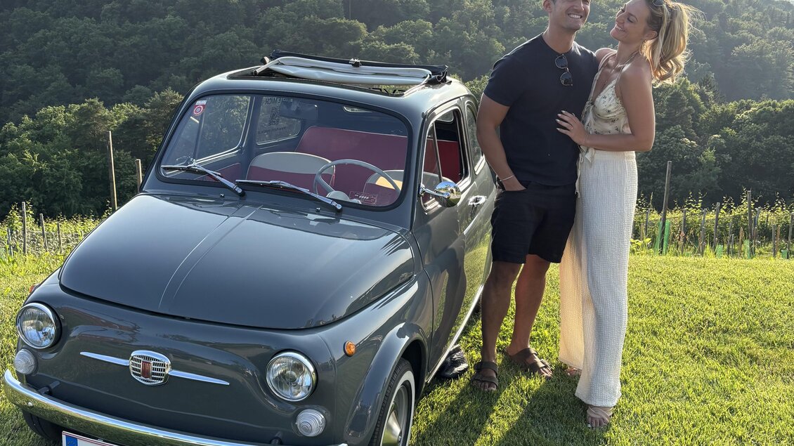 Ein Paar posiert neben einem vintage Auto auf einer grünen Wiese. Im Hintergrund sieht man sanfte Hügel und Weinreben.