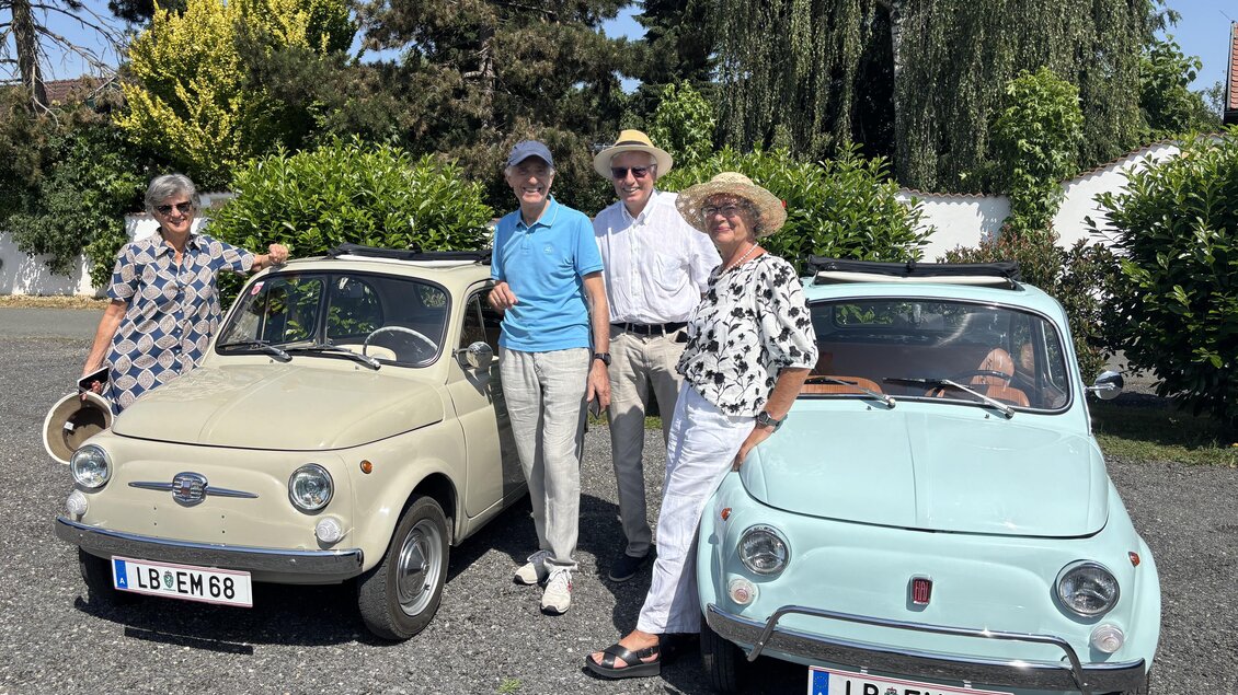 Vier Personen stehen lächelnd neben zwei klassischen Autos auf einem Kiesplatz. Die Umgebung ist grün und sonnig, was eine entspannte Atmosphäre schafft.